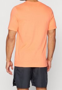 Mies, jolla on päällään yksinkertainen oranssi lyhythihainen t-paita ja mustat urheilushortsit, kuvattu selkäpuolelta tasaisella taustalla.