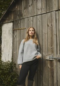 Pulover tricotat în albastru deschis și crem, cu dungi diagonale, asortat cu pantaloni negri și cizme maro până la genunchi; fundal din lemn texturat.