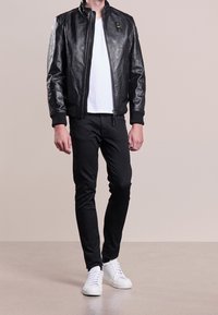 Veste bomber en cuir noir avec fermeture éclair et poignets à côtes, portée sur un t-shirt blanc, associée à un pantalon noir et des baskets blanches.