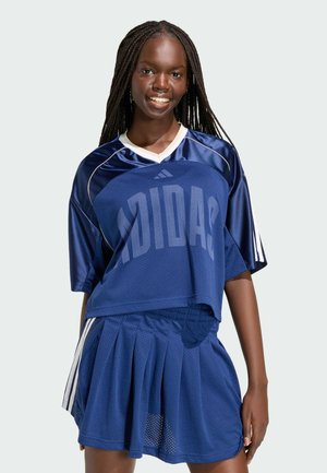 Usměvavá žena s dlouhými copy, oblečená v modrém sportovním dresu Adidas a plisované sukni s bílými pruhy na rukávech a bocích.