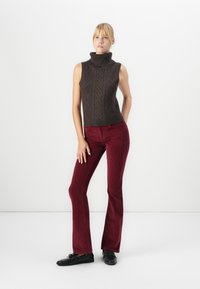 Nő áll sleeveless sötétbarna garbó pulóverben, bordó kordbársony nadrágban és fekete fűzős cipőben, fehér háttér előtt.