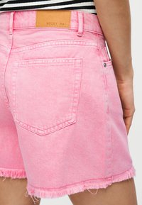 Άτομο που φοράει ροζ τζιν σορτς με ξεφτισμένο τελείωμα και καφετί δερμάτινη ετικέτα επωνυμίας στη μέση, σε συνδυασμό με μαύρη και λευκή ριγέ μπλούζα.