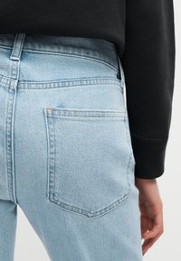 Άτομο που φοράει ανοιχτό μπλε τζιν με ορατή πίσω τσέπη και μαύρο μπλουζάκι με μακριά μανίκια, χέρι χαλαρό στο πλάι.