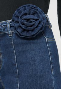 Τα τζιν από τζιν υλικό έχουν μια γαλάζια υφασμάτινη διακόσμηση με λουλούδια στη μέση. Τα τζιν είναι εφαρμοστά, με ομαλή υφή και εμφανείς ραφές.