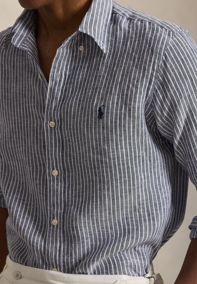 Άνδρας που φορά μπλε και λευκό πουκάμισο μ ε κάθετες ρίγες, κουμπωμένο μέσα σε λευκό παντελόνι, με σκούρο κεντημένο λογότυπο στο στήθος.