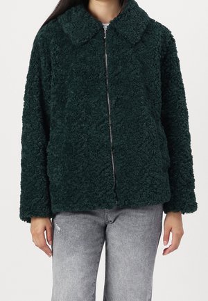 Veste en polaire texturée vert foncé avec col et fermeture éclair à l'avant, portée avec un jean gris clair.