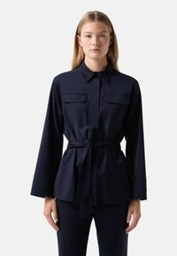 Navy blue shirt jacket made of smooth fabric, featuring a collar, two chest pockets, and a waist tie for adjustable fitting.