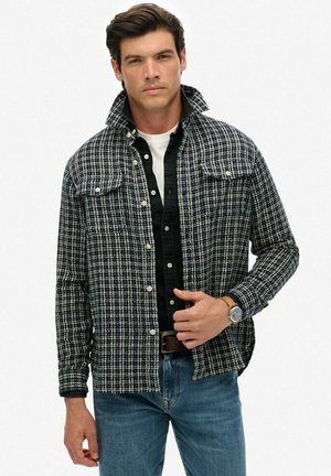 Man in een zwart-wit geruite overhemden met knopen over een zwart overhemd en wit T-shirt, gecombineerd met blauwe jeans en een polshorloge.