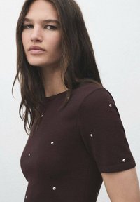 Camicia marrone a maniche corte con decorazioni in argento, forma aderente, texture liscia, collo giro, leggero luccichio e maniche arrotolate.