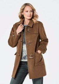 Brauner Wollmischungs-Mantel mit hohem Kragen, Knopfverschlüssen, seitlichen Taschen und Knopfdetails. Das Modell trägt ein blau gemustertes Oberteil darunter.