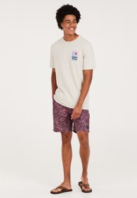 Light beige cotton t-shirt with a chest graphic and pink and black leaf-patterned swim shorts. Model wears brown flip-flops.