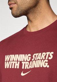 Κόκκινο βαμβακερό t-shirt με μεγάλο μπεζ κείμενο «Η ΝΙΚΗ ΞΕΚΙΝΑ �ΜΕ ΤΗΝ ΠΡΟΠΟΝΗΣΗ» και το λογότυπο της Nike κάτω από το κείμενο. Κλασικός σχεδιασμός με στρογγυλή λαιμόκοψη.