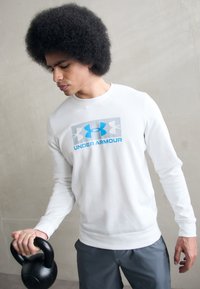 Sudadera blanca de manga larga con un diseño de logo "Under Armour" en azul y gris, combinada con shorts deportivos grises. Sosteniendo un kettlebell negro.