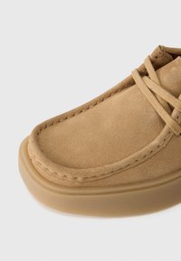 Mocassin en daim marron clair avec détail de la pointe cousue et lacets assortis sur un fond neutre.