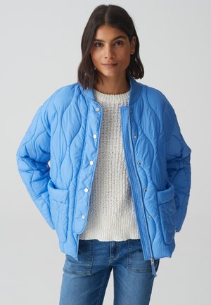 Lichtblauwe gewatteerde jas met een ritssluiting aan de voorkant, drukknoopsluitingen en twee voorkwapen, gecombineerd met een gestructureerde crèmekleurige trui en jeans.
