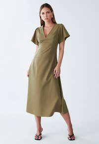Ženska oblečena v olivno zeleno midi obleko z V-izrezom in kratkimi flitrastimi rokavi, v kombinaciji s črnimi sandali na peto s trakci, na bež ozadju.