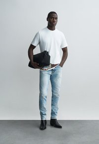 Uomo con maglietta bianca, jeans azzurri, stivali neri, che tiene in mano una giacca nera piegata, in piedi davanti a una parete semplice di colore grigio chiaro.