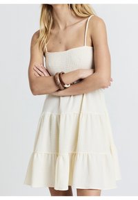 Robe à volants de couleur crème claire avec un corsage smocké, des bretelles fines et un tissu texturé. Soulignée par un ourlet fluide.