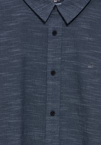 Camicia button-up blu scuro con tessuto testurizzato, dotata di colletto, sei bottoni e un piccolo dettaglio del logo vicino alla parte inferiore destra.