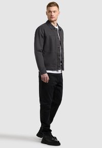Homme portant une veste noire à boutons, une chemise blanche, un pantalon noir et des chaussures noires, debout et regardant sur le côté sur un fond uni.