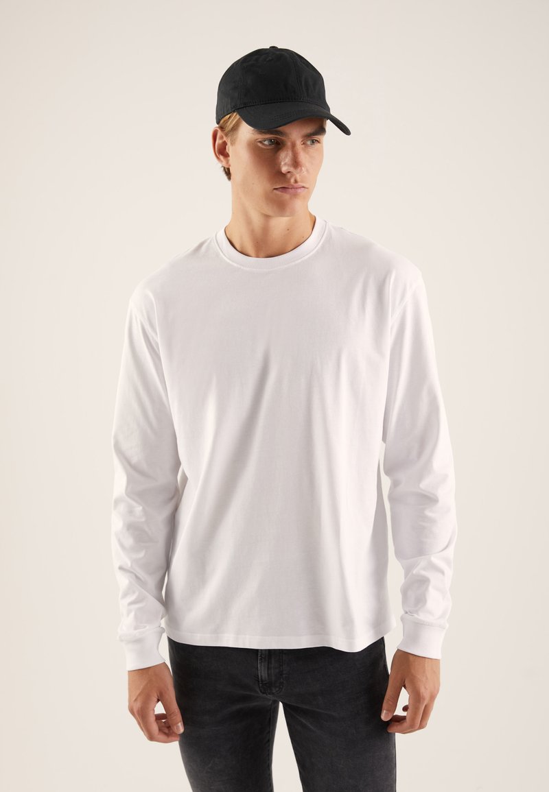 Weißes Langarmshirt mit Rundhalsausschnitt, kombiniert mit einer schwarzen Mütze. Das Shirt hat eine glatte Textur und eine lockere Passform.