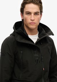 Giacca nera impermeabile con colletto alto, tasche con zip e dettagli del logo discreti. Presenta una finitura opaca e un cappuccio regolabile.