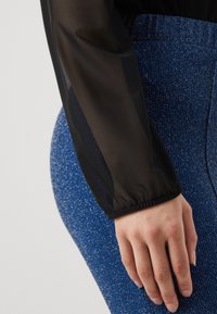Svart, gjennomsiktig langermet topp med elastiske mansjetter ved siden av en hånd, kombinert med blå, teksturerte bukser med et skinnende mønster.