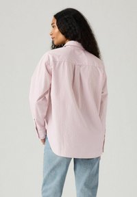 Camicia a righe rosa e bianche con maniche lunghe, taglio morbido e dettaglio di piega sul retro, abbinata a jeans azzurri chiari.