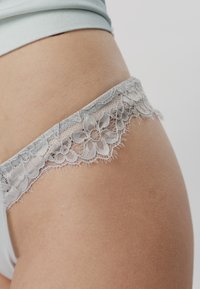 Κοντινό πλάνο ενός ισχίου που φοράει λευκά εσώρουχα με δαντέλα και ανοιχτόχρωμο τοπ σε απλό φόντο.