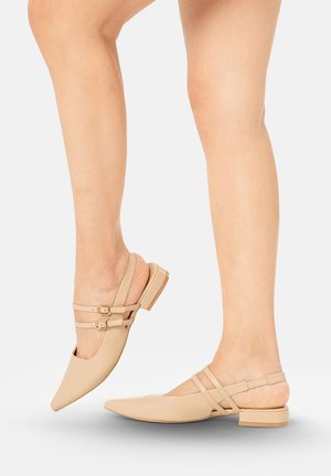 Benen met beige puntige slingback platte schoenen met dubbele gespbanden, één voet opgetild, tegen een witte achtergrond.
