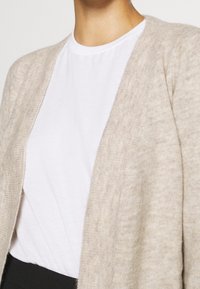 Beige knit cardigan with an open front design, featuring a soft texture. Worn over a white t-shirt, suitable for layering.