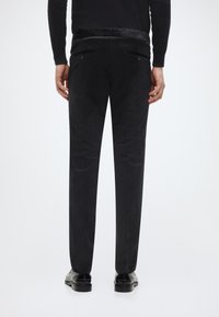 Schwarze, maßgeschneiderte Hose mit glatter Textur, geradem Beinverlauf, zwei Gesäßtaschen und dezentem Glanz. Getragen mit schwarzen Schuhen.