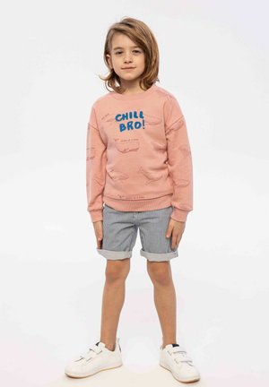 Sweatshirt rose avec motifs de baleines et texte bleu "CHILL BRO!", associé à un short rayé et des baskets blanches. Design décontracté et confortable.