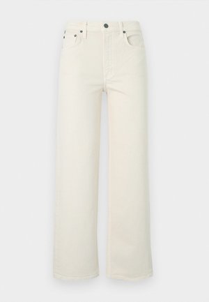 Nő fekete, szakadt szegélyű V-nyakú felsőben, törtfehér, bő szárú nadrágban fekete övvel, fekete magassarkú szandálban és sötét napszemüvegben, állva.