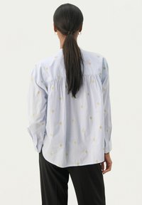 Camicia azzurro chiaro con motivi a diamante dorati, maniche lunghe, design raccolto sul retro e orlo arrotondato. Abbinata a pantaloni neri.