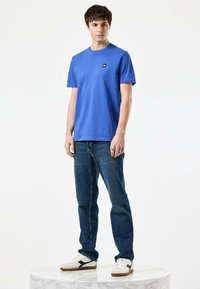 Blå crew-neck t-skjorte, kombinert med mørk denim jeans. Modellen har på seg hvite sneakers med svarte detaljer. Enkel, uformell design.