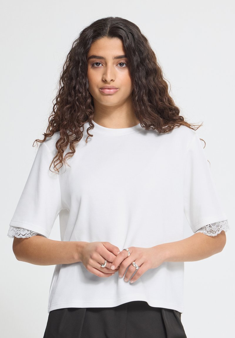 Jeune femme aux longs cheveux bouclés portant une chemise blanche à manches courtes avec dentelle et un pantalon noir, mains jointes avec des bagues aux doigts.