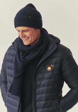 Tumši zilas ribas adīta cepure un šalle papildina tumšu piedurkņu jaku ar lāča uzaplīšu, kas ir ar kviltētu dizainu un fakturētu audumu.