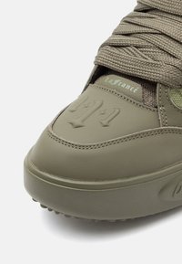 Olivzaļas krāsas sporta apavi ar strukturētu ādu, izceļamu logotipa detaļu un plakaniem auklām. Raksturo gluds dizains, polsterēta zole un šuvju akcenti.