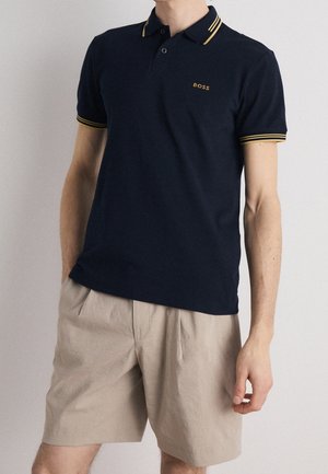 Mand iført en navy polo med gule striber på kraven og ærmerne og beige plisserede shorts, stående med den ene hånd i lommen.