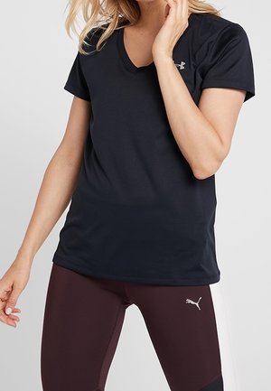 Frau trägt ein schwarzes Under Armour V-Ausschnitt T-Shirt und kastanienbraune Puma-Leggings, posiert mit einer Hand nahe dem Gesicht vor einem schlichten Hintergrund.
