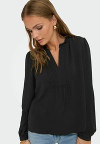 Svart långärmad blus med V-ringning, texturerat tyg, volangkrage och dekorativa veck, ihopparad med ljusblå jeans.