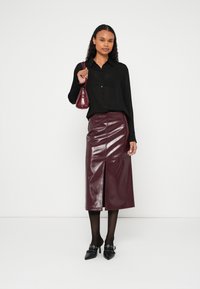 Melna krojuma krekls, burgundijas krāsas lakādas midi svārki ar priekšējo šķēlumu, caurspīdīgas melnas zeķubikses un melni norādīti augstpapēžu apavi ar metāla detaļām.