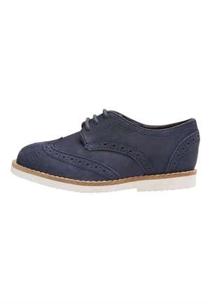 Navy blå læder oxford sko med brogue-detaljer, hvid sål, beige kant og snørelukning, vist fra siden.