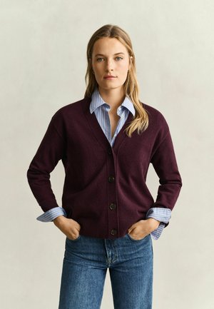 Femme debout les mains dans les poches, portant un cardigan boutonné violet foncé par-dessus une chemise rayée bleu clair et un jean bleu sur un fond uni.