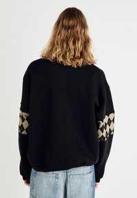 Sweat-shirt noir à coupe décontractée, avec des accents texturés à motif losange beige et noir sur les manches. Tissu doux, ourlet côtelé.