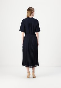 Navy pleated dress with short sleeves and a belted waist, featuring a back pleat detail and a flowy skirt. Worn with clear heeled shoes.