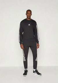 Crni pamuk sweatshirt s bijelim prugama na rukavima, s malim logom. Uparen s odgovarajućim crnim hlačama, također s bijelim prugama.