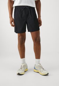 Zwarte sportbroek met een elastische tailleband en trekkoord, gladde textuur en een pasvorm tot de knie, gecombineerd met lichtgekleurde hardloopschoenen.