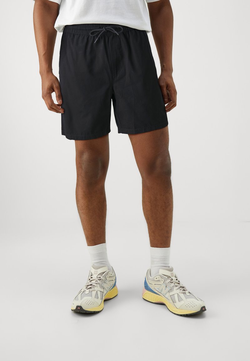 Zwarte sportbroek met een elastische tailleband en trekkoord, gladde textuur en een pasvorm tot de knie, gecombineerd met lichtgekleurde hardloopschoenen.